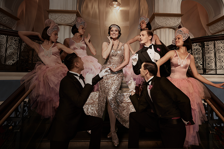 Singer Julie Lea Goodwin stands on a large staircase wearing a gold dress, surrounded by dancers in formal wear