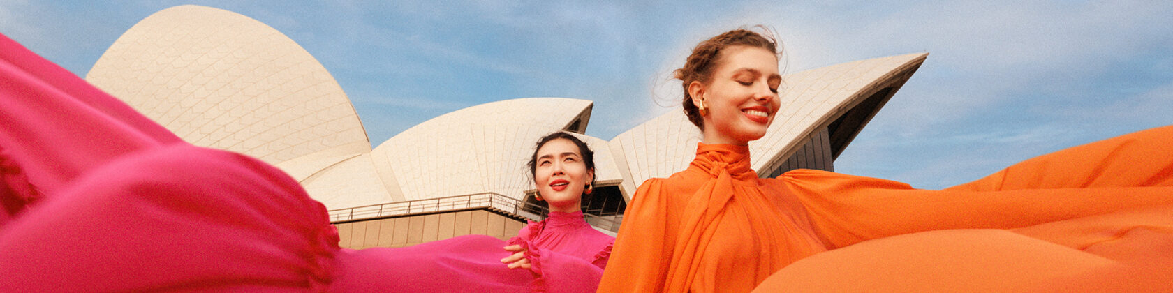 Two young women in beautiful flowing colourful dresses in front of the Sydney Opera House on a summer's day.