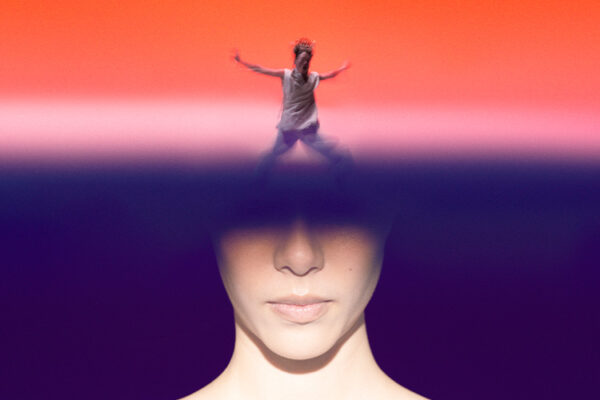 a close-up of a performer’s face is framed against a deep purple background, with a horizontal band obscuring the eyes. Above this band, a smaller figure stands with arms outstretched against a vivid red backdrop. The stylised staging and strong colour contrast create a dramatic, surreal atmosphere.